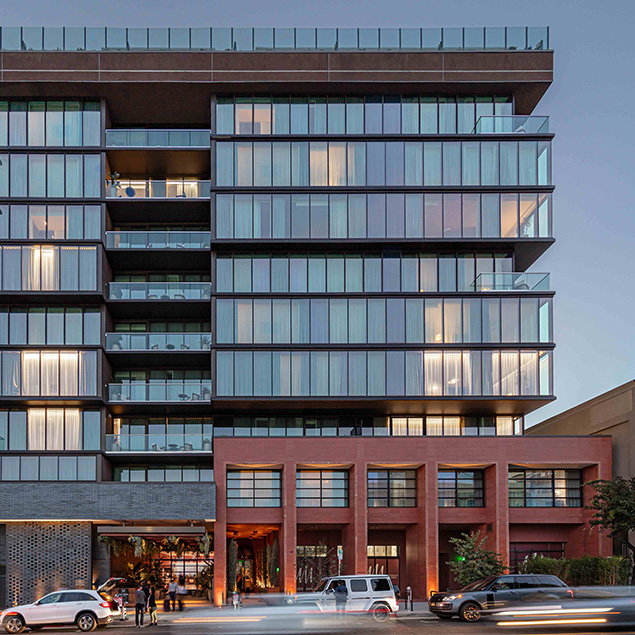 A modern multi-story building with large glass windows and balconies, featuring a lower section with brick and a main entrance. Several cars are parked and driving in front of the building at dusk.