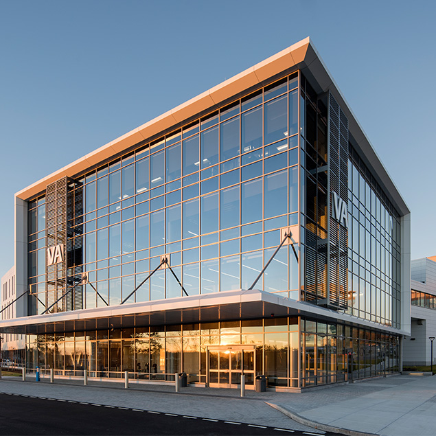 A modern glass building with VA logos on its facade, reflecting the sunset sky. The structure features large windows, an angled roof, and a covered entrance.