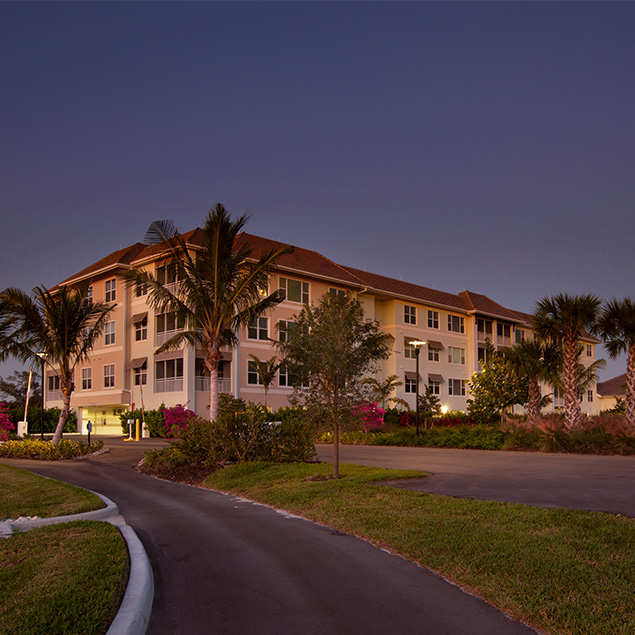 A four-story apartment building with warm exterior lighting stands amid palm trees and landscaped greenery under a deep blue evening sky. A curved driveway leads to the entrance.