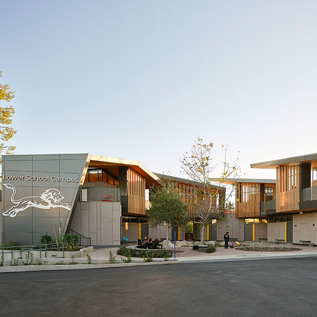 Modern school building with large windows and wooden accents, a logo of a lion on the facade, and landscaped outdoor areas with trees and benches. A few people are visible near the entrance.