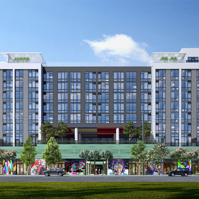 A modern mid-rise building with many windows, rooftop terraces, and colorful murals at the street level. Trees and cars line the front, and a grassy area is visible in the foreground under a blue sky.