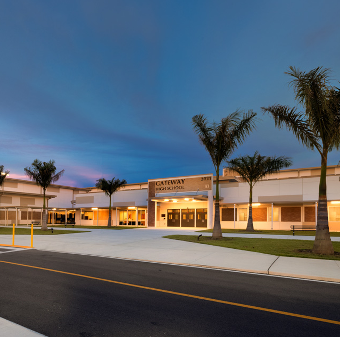 A modern high school building with palm trees lining the entrance, illuminated by exterior lights at dusk under a colorful sky. A driveway and crosswalk are visible in the foreground.