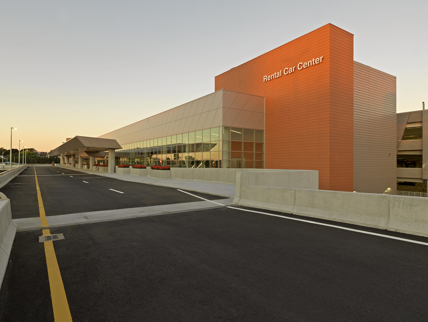 A modern, multi-story Rental Car Center building with large glass windows and orange panels, next to an empty asphalt road, photographed at sunset.