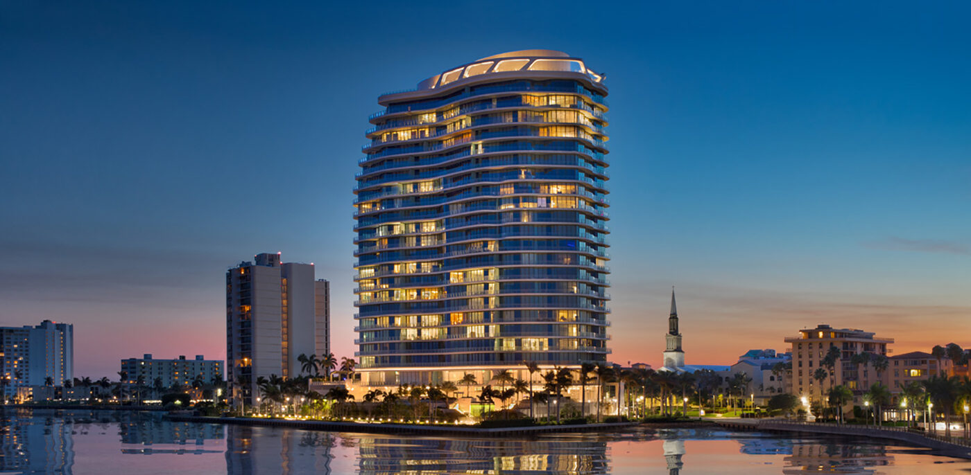 A modern, multi-story waterfront building is illuminated at dusk, reflecting in the calm water below, with nearby palm trees, surrounding buildings, and a church steeple visible against a colorful evening sky.