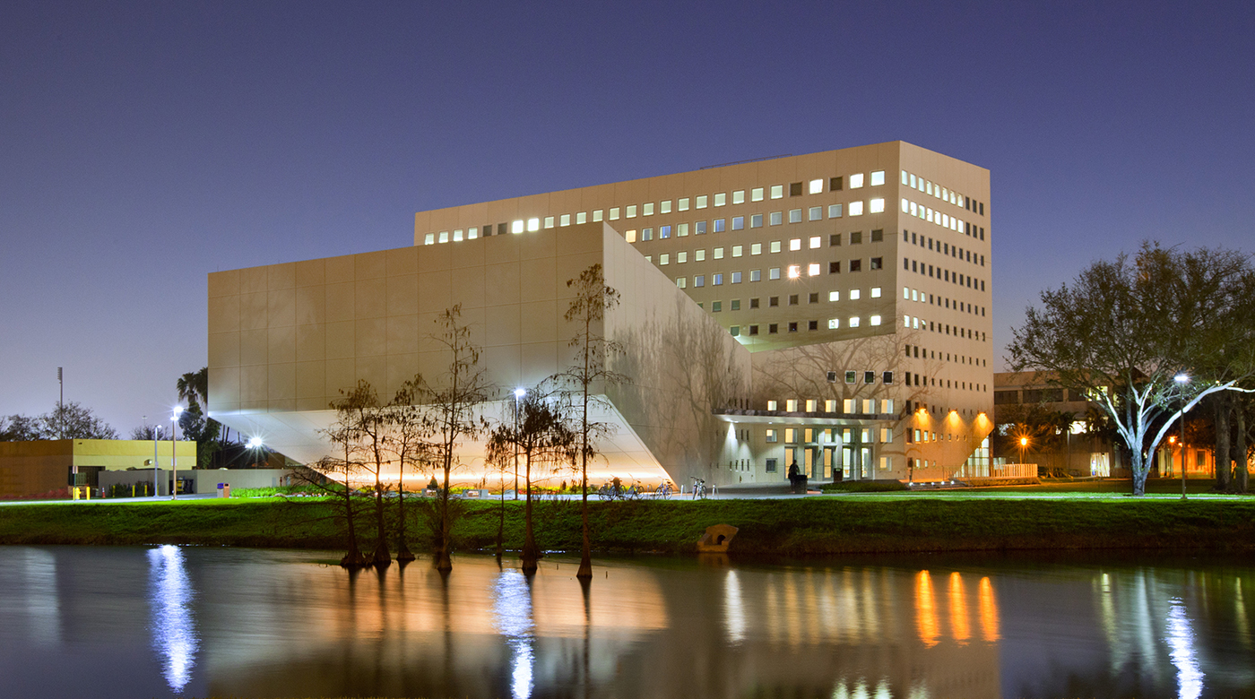 A modern, angular building with many lit windows is reflected in a calm river at night. Trees line the grassy riverbank, and outdoor lights illuminate the structure and surrounding area.