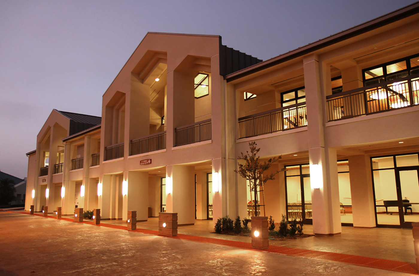 A modern two-story building with large windows and bright exterior lights at dusk, featuring columns and a spacious walkway in front.