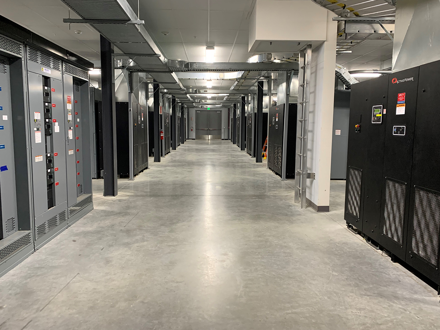 A clean, modern data center with rows of black server racks and electrical panels lining both sides of a wide hallway under a ceiling with exposed cables and pipes.