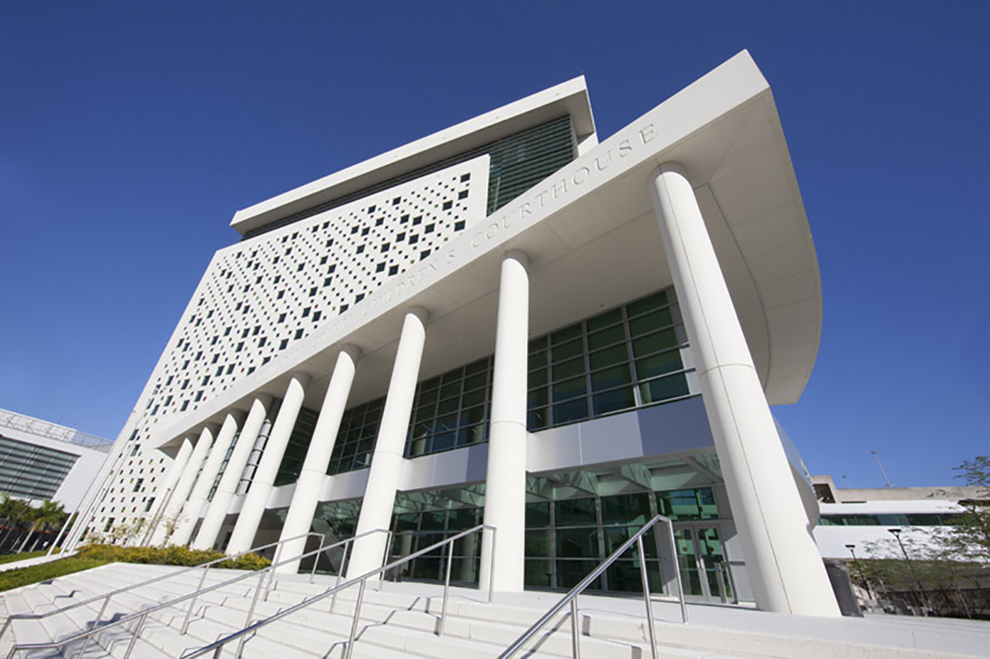 A modern courthouse building with tall white columns, geometric patterned facade, glass windows, and staircases leading to the entrance against a clear blue sky.