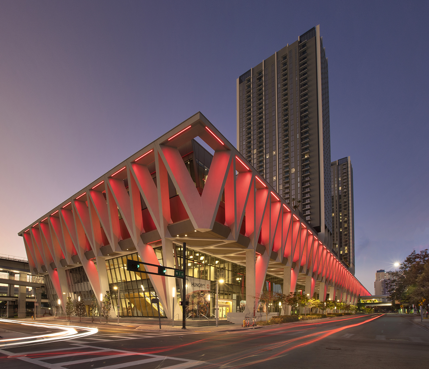 A modern building with striking V-shaped columns is illuminated with red lights at dusk. Behind it, a tall skyscraper rises into the twilight sky. Light trails from passing vehicles are visible on the street in front.