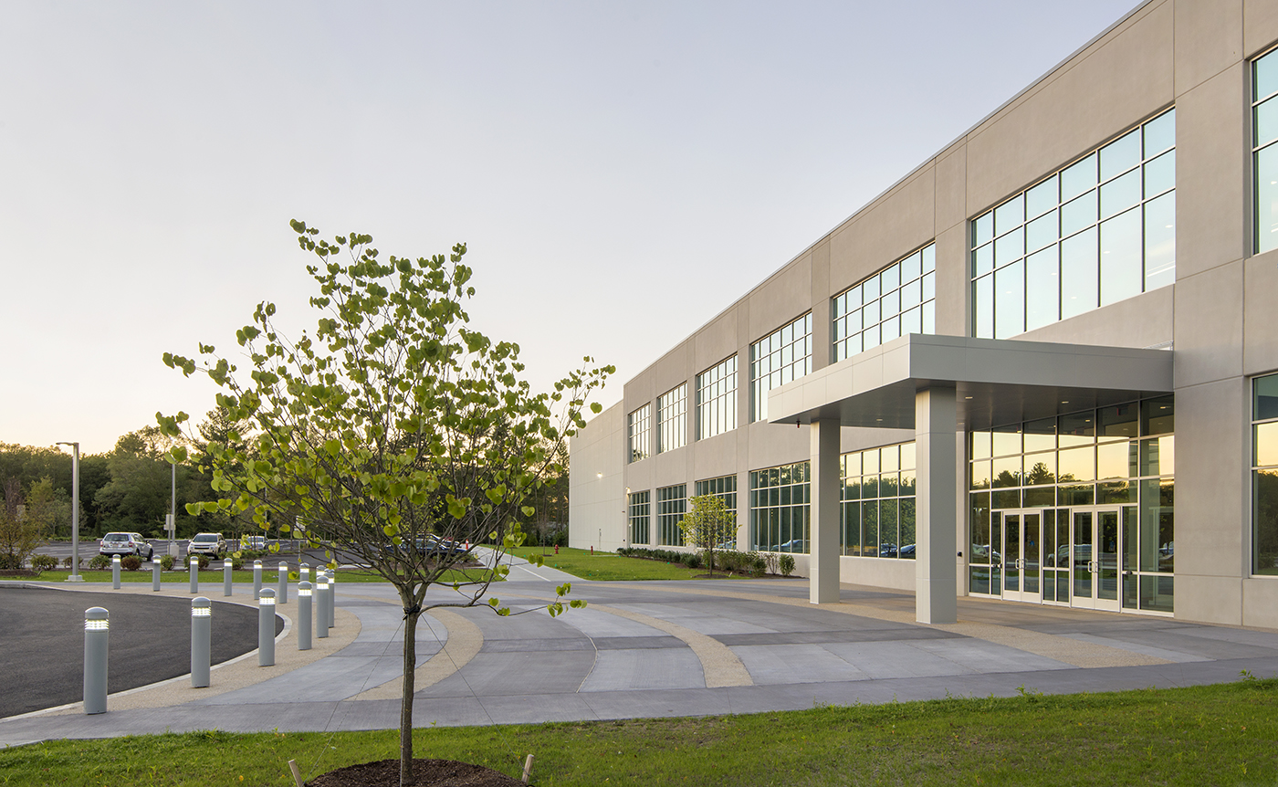 Modern office building with large windows, a main entrance with an awning, and a small tree planted near a clean, curved sidewalk. Cars are parked in the lot, and the surroundings are green with trees.