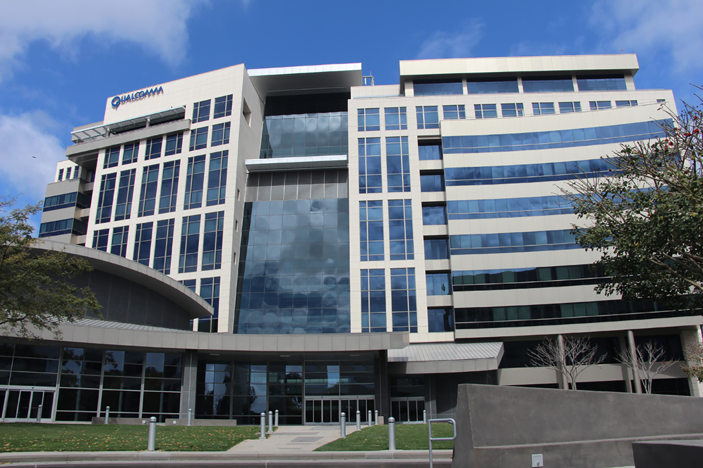 A modern multi-story office building with large glass windows and Qualcomm signage on top, set against a blue sky with scattered clouds and surrounded by trees and grass.