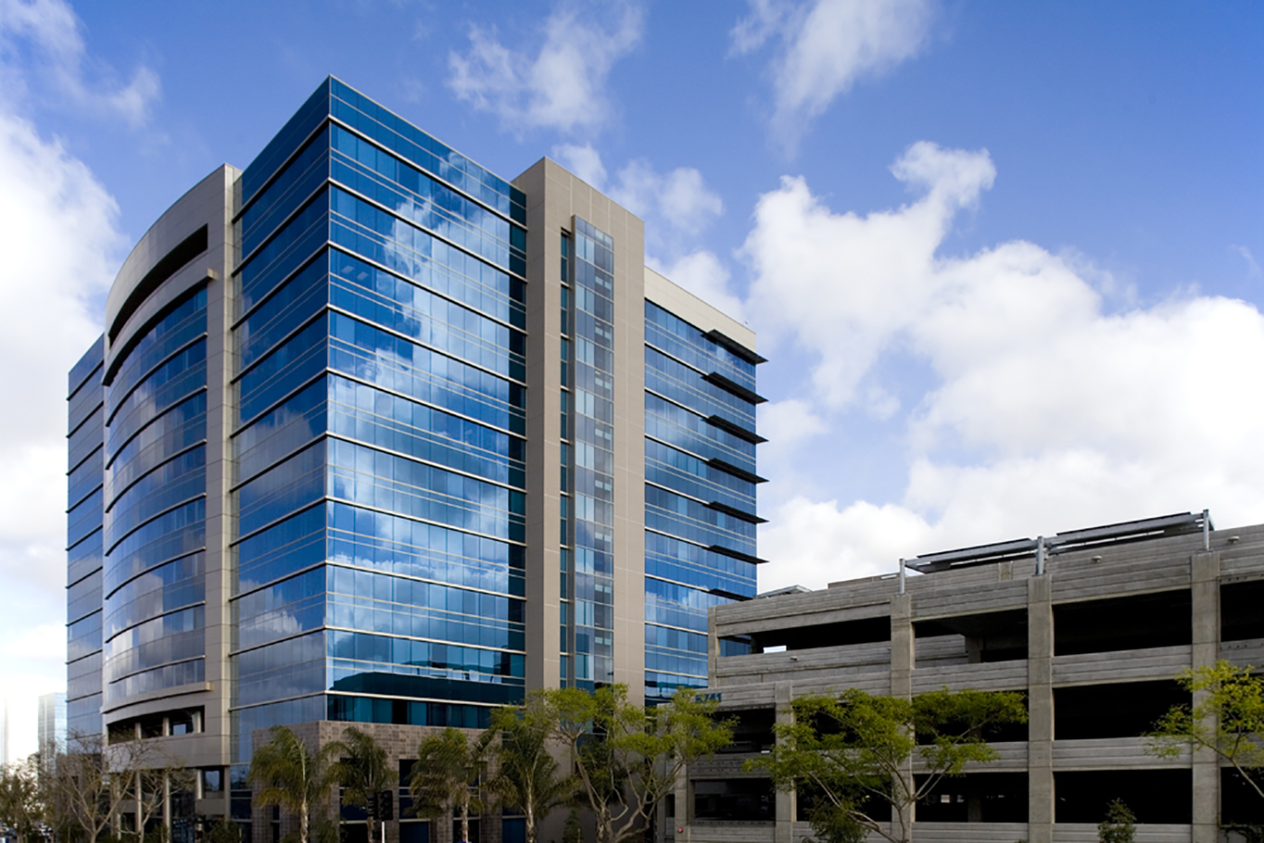 A modern glass office building with blue-tinted windows stands next to a concrete parking garage. Palm trees line the front, and the sky is partly cloudy.