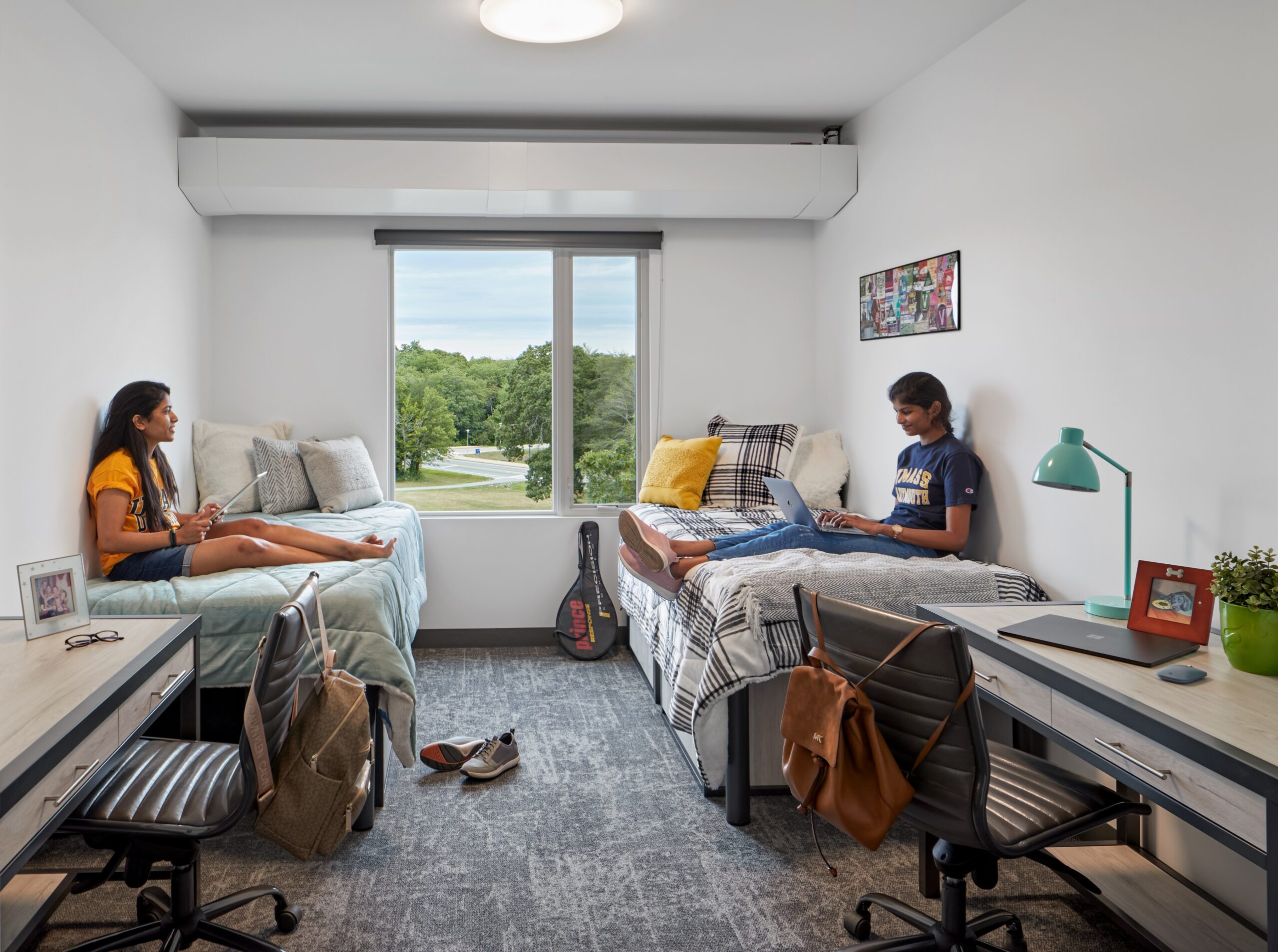 Two students sit on separate beds in a bright Higher Education dorm room with desks, chairs, and a window overlooking greenery. One uses a laptop while the other looks out the window. The tidy rooms are decorated with personal items.