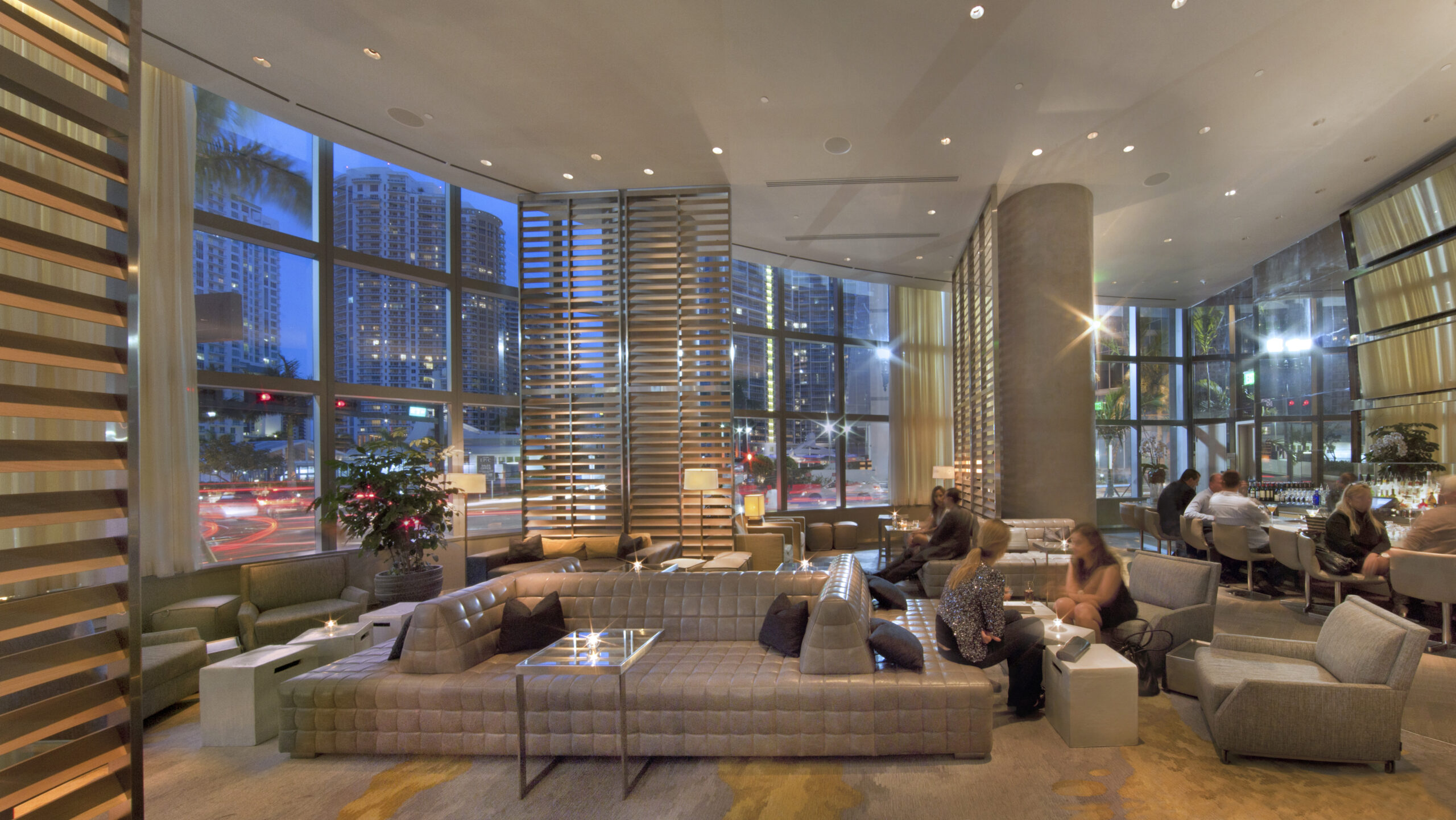 A modern hotel lounge with plush seating, floor-to-ceiling windows, and city views at dusk. People are relaxing on sofas and at the bar, enjoying warm lighting and a cozy, elegant atmosphere that reflects true hospitality.