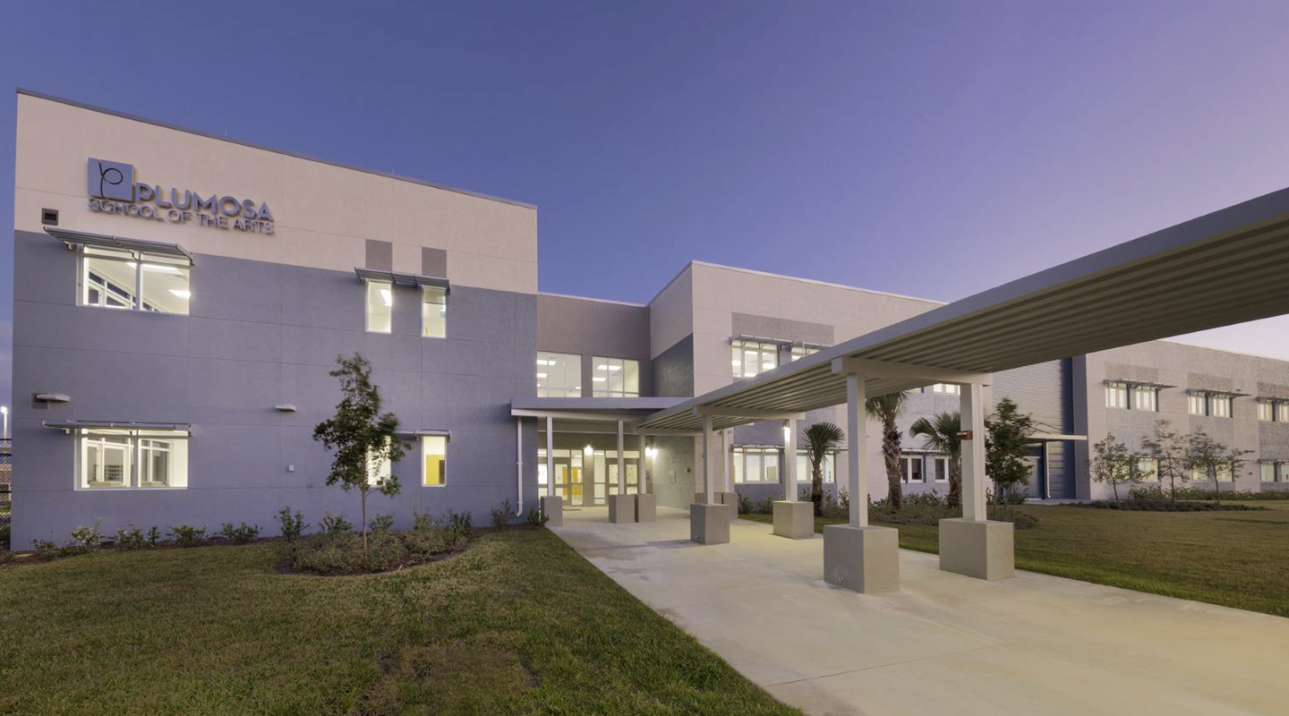 A modern two-story school building labeled Plumosa School of the Arts, with large windows, a covered walkway, and landscaped grounds, photographed at dusk with lights on inside.