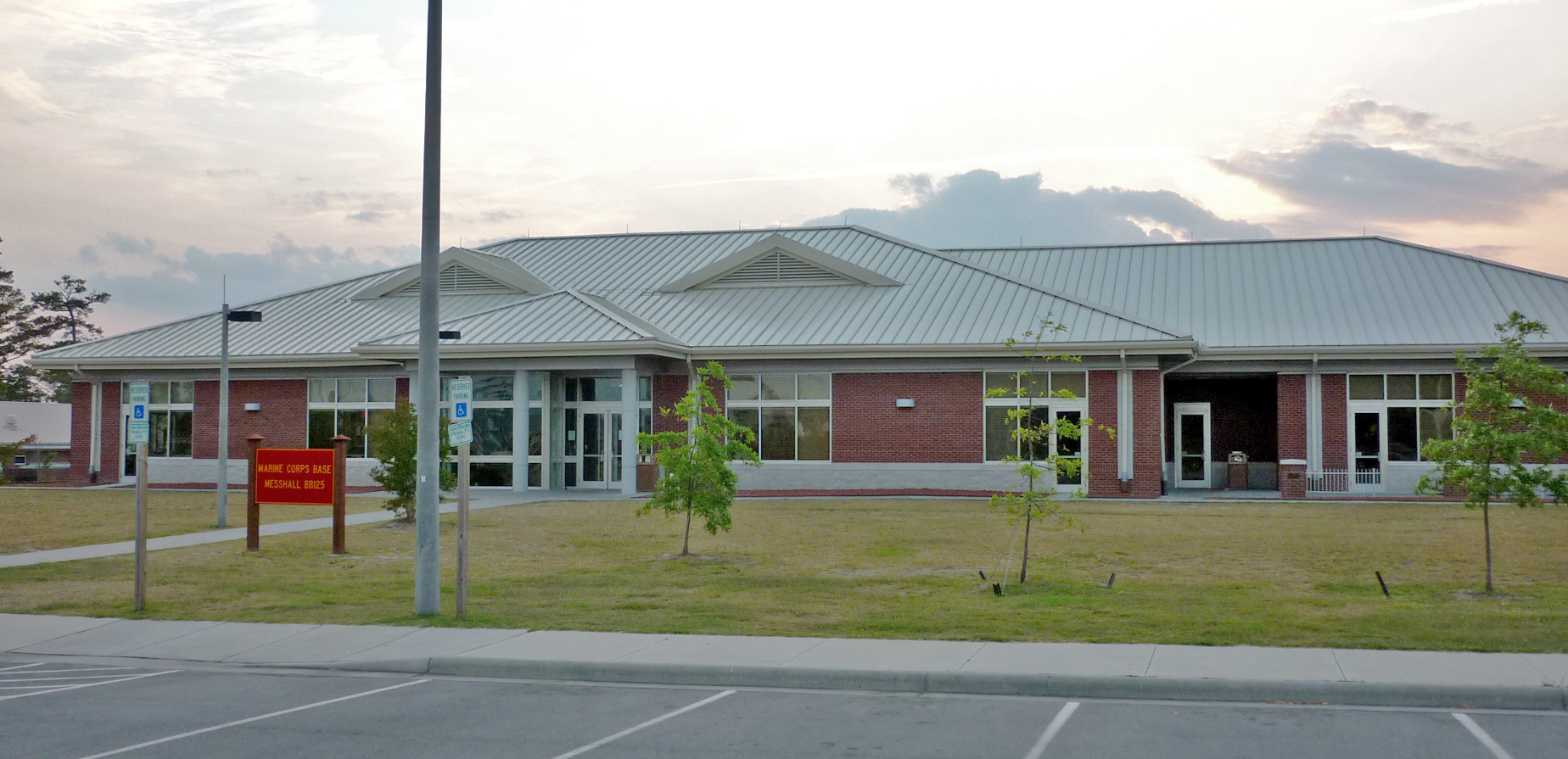 A single-story brick building with a metal roof, large windows, and a small lawn with young trees in front. Theres a red sign near the entrance; the sky is partly cloudy at sunset.