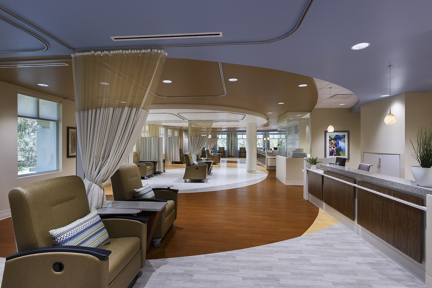 A spacious, modern medical infusion center with comfortable reclining chairs, privacy curtains, wood and light gray flooring, and a reception desk, all under soft lighting with large windows providing natural light.