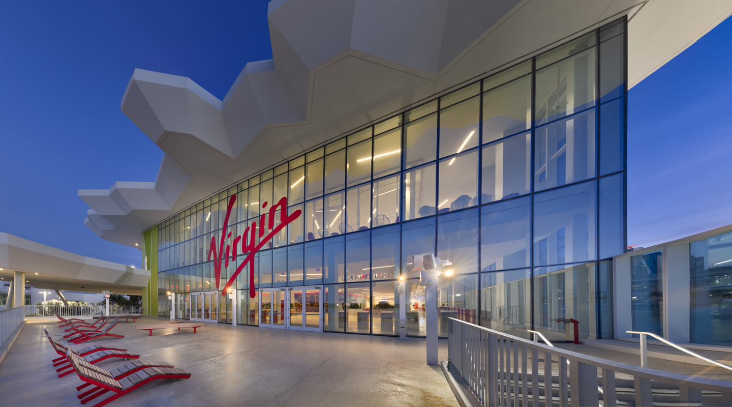 A modern building with large glass windows displaying a bright red Virgin logo. The entrance is flanked by red benches and illuminated by exterior lights, with an overhanging geometric white roof.