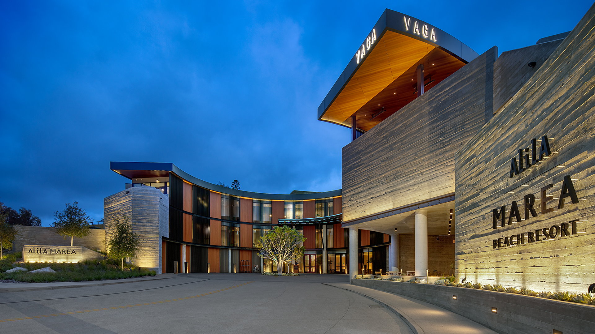 Modern beachfront resort with curved architecture, large windows, and wood accents illuminated in the evening; signs read “ALILA MAREA BEACH RESORT” and “VAGA” above the entrance. A tree and driveway are in the foreground.