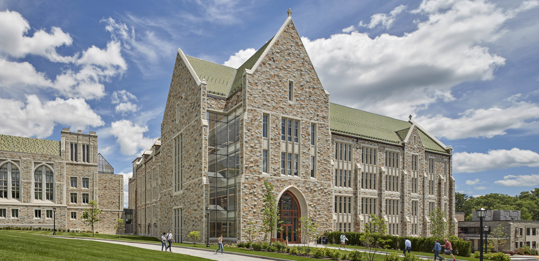 A large stone university building with Gothic architecture, tall windows, and a green roof, sits under a partly cloudy sky. People walk on paths and grass in front of the building.