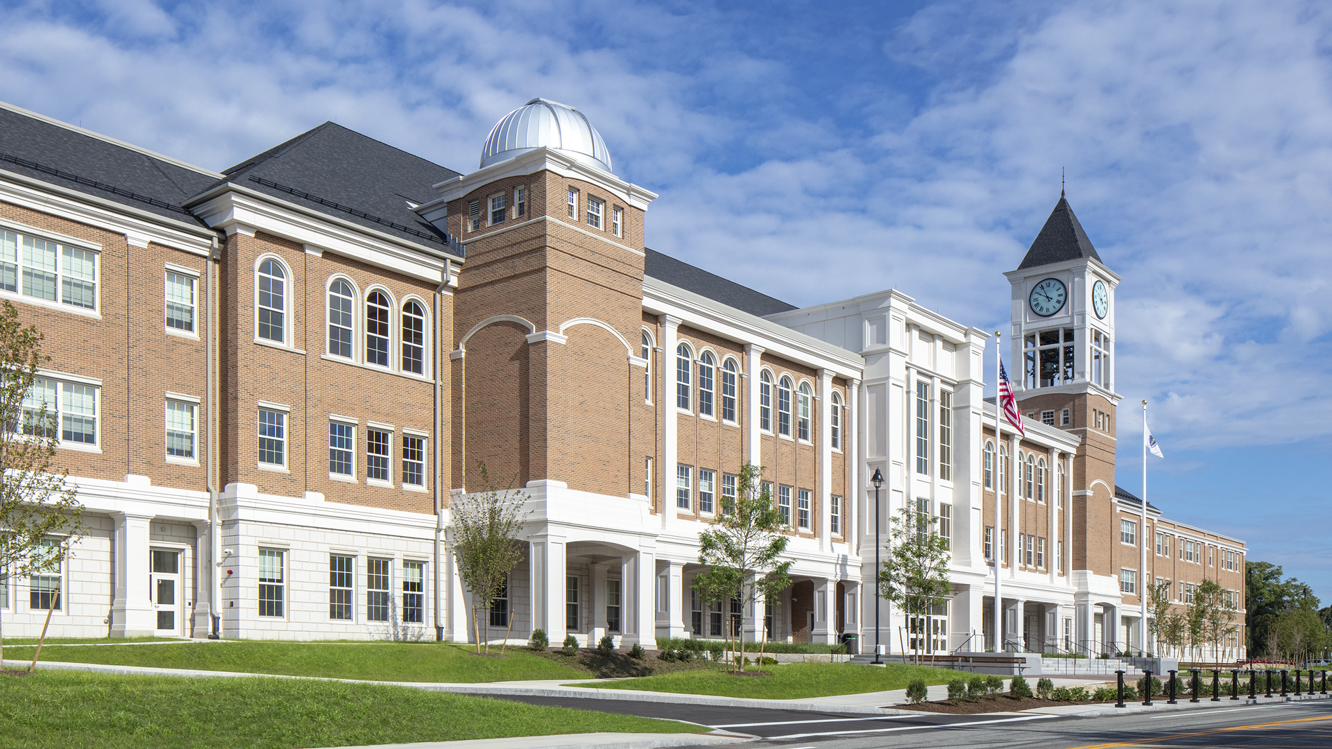 A large, modern brick school building with a clock tower and dome under a blue sky; American flags hang near the entrance, and landscaped greenery surrounds the front.
