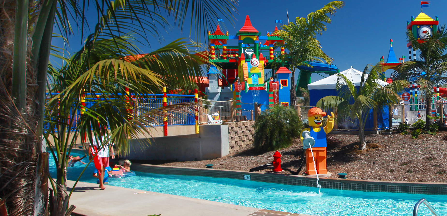 A colorful LEGO-themed water park features a lazy river, palm trees, bright LEGO sculptures, and a vibrant play structure under a clear blue sky. Children and adults enjoy the water attractions.