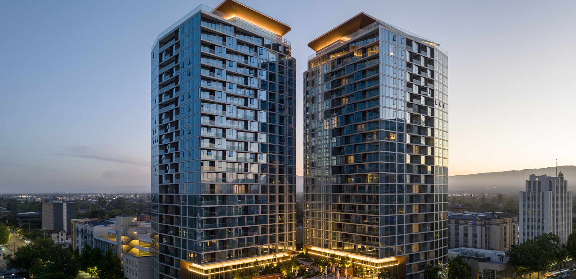 Two modern high-rise apartment buildings with glass exteriors are illuminated at dusk, standing side by side above a cityscape with trees and surrounding lower buildings.