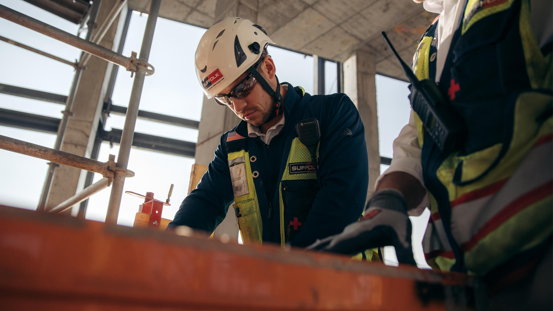 A construction worker in a helmet and safety gear focuses on a task, leaning over an orange surface, as teamwork thrives across different sectors at a busy building site.