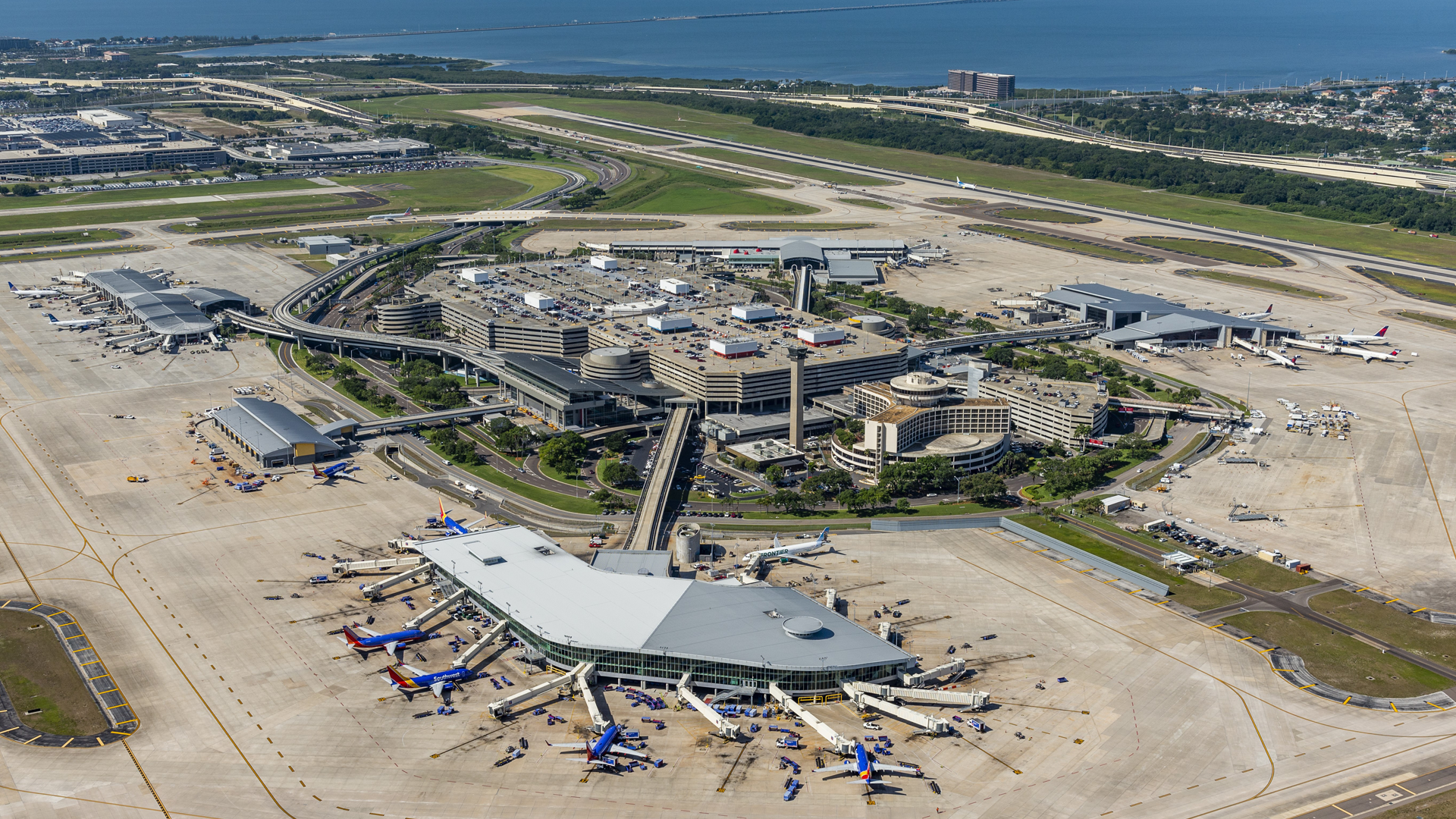 Aerial view of a large airport with multiple terminals, airplanes parked at gates, runways, taxiways, and surrounding roads, with water and residential areas in the background.