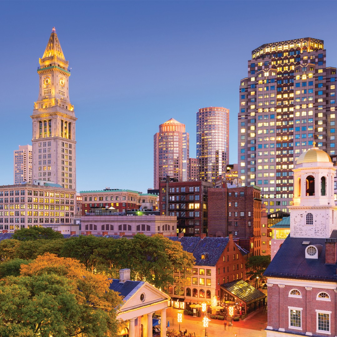 Boston city skyline at dusk, featuring illuminated historic and modern buildings, including the Custom House Tower, with trees and streetlights in the foreground under a clear sky.