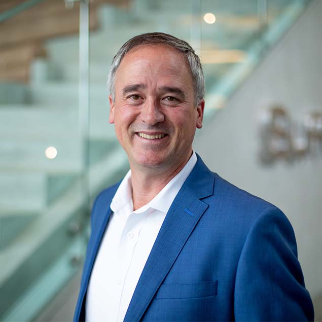 A middle-aged man in a blue suit and white shirt smiles at the camera, standing in front of glass railings and a modern staircase in an office building.