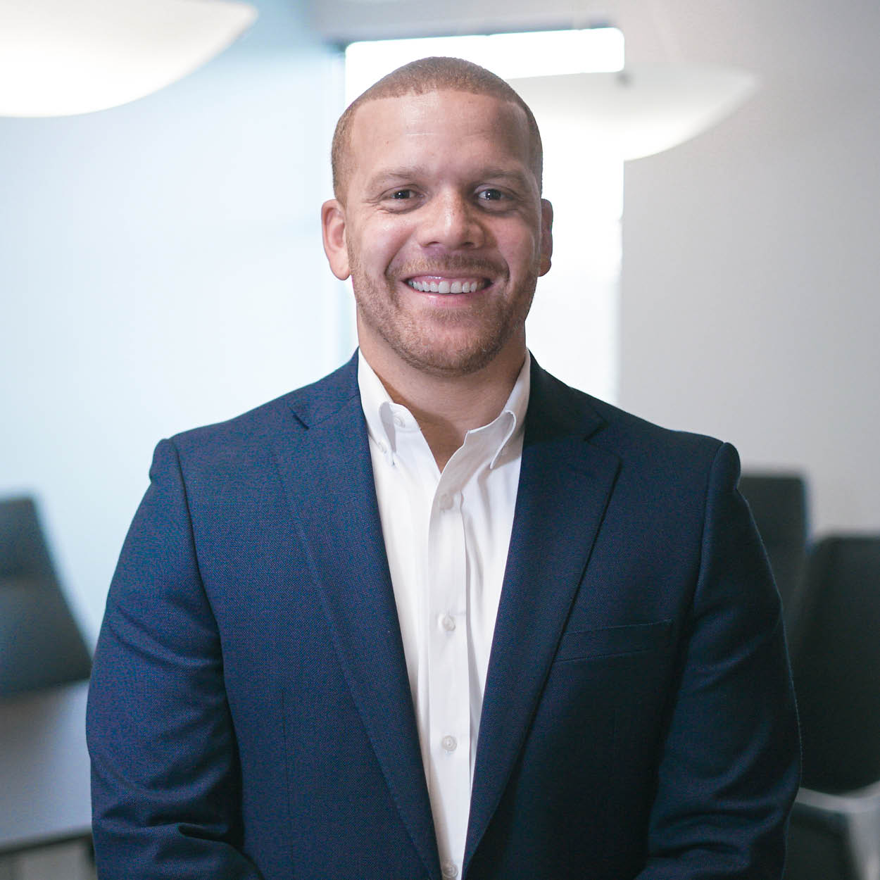 A man in a navy blue suit and white shirt stands smiling in a modern office with soft lighting and blurred chairs in the background.