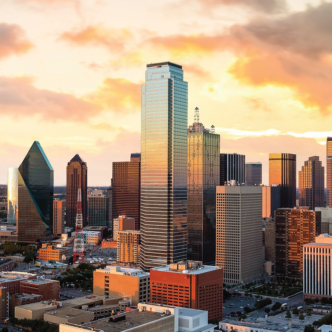 Downtown Dallas skyline at sunset, featuring tall modern skyscrapers, including the glass-covered Fountain Place and other high-rise buildings, under a sky with orange-tinted clouds.