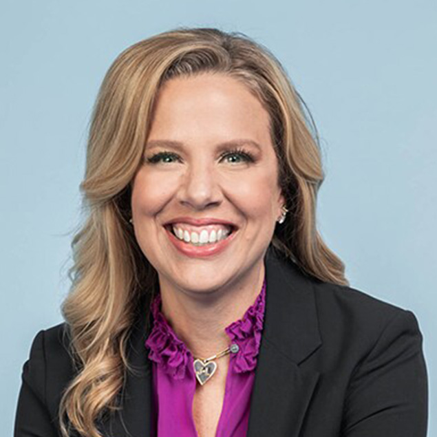 A woman with wavy blonde hair wearing a black blazer over a purple blouse and a heart-shaped necklace smiles in front of a plain light blue background, representing the Advisory Board.