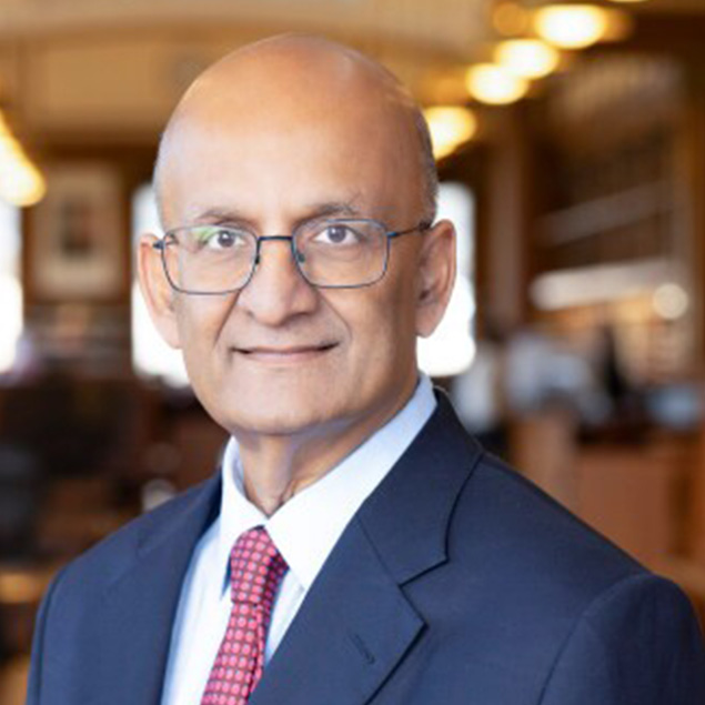 A bald man wearing glasses, a navy suit, white shirt, and red tie, smiling slightly in a warmly lit indoor setting with blurred lights and shelves—he exudes the confidence of an Advisory Board member.