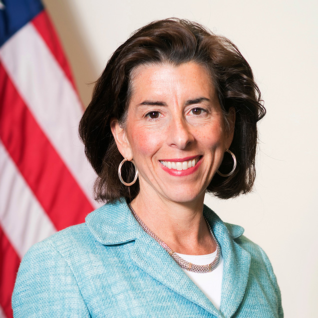 A woman with short brown hair, wearing a light blue blazer, white top, hoop earrings, and a necklace, smiles in front of an out-of-focus American flag and light background—perfect for an Advisory Board introduction.