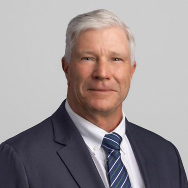 A middle-aged man with short gray hair, part of an Advisory Board, wearing a dark suit, white dress shirt, and blue striped tie, posed against a plain light gray background.