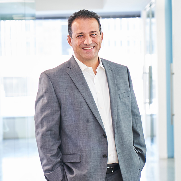 A man in a grey suit and white shirt stands smiling in a bright modern office hallway with large windows, ready to collaborate with his team.