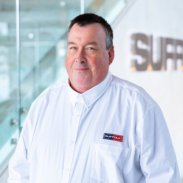 A man with short dark hair smiles slightly, wearing a white button-up shirt with a SUFFOLK logo on the chest. He stands indoors near glass railings and a blurred SUFFOLK sign, representing pride in his Team.