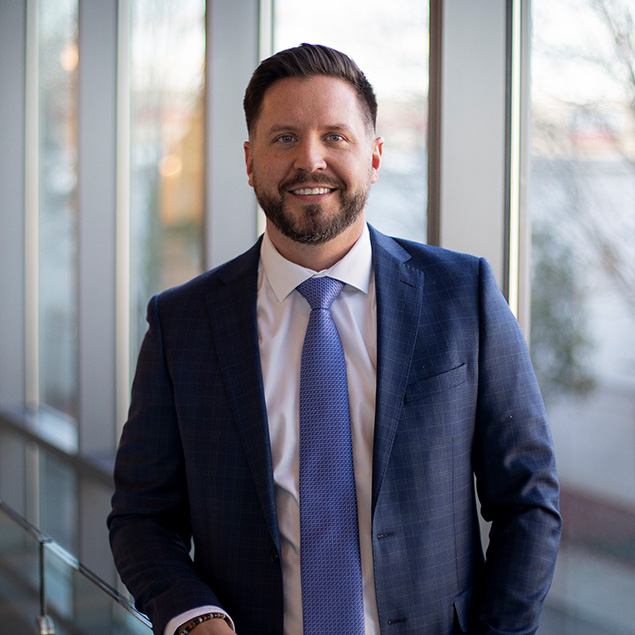 A man with short brown hair and a beard, wearing a blue suit and light blue tie, stands smiling by large windows with blurred trees outside.