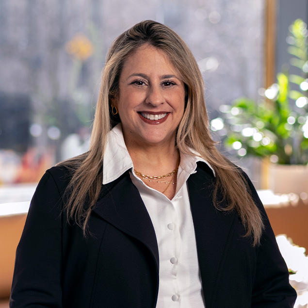 A woman with long blonde hair, wearing a white shirt and black blazer, smiling while standing indoors in front of a window with blurred greenery and soft sunlight in the background.