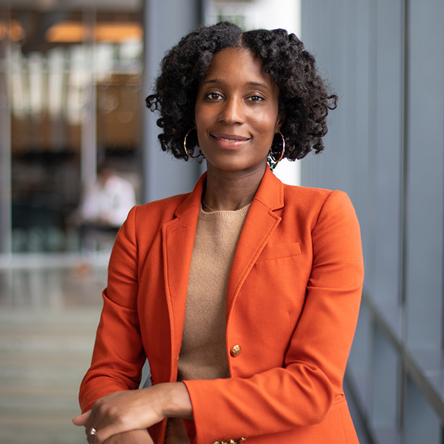 A woman with curly hair wearing a bright orange blazer and tan top stands indoors, smiling confidently with arms crossed, in front of glass panels and a blurred office background.