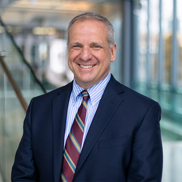 A middle-aged man in a navy suit, striped dress shirt, and multicolored striped tie smiles confidently, embodying executive leadership in a bright, modern indoor setting with glass and blurred background elements.