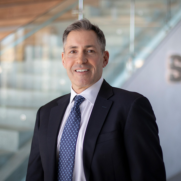 A man in a dark suit and patterned tie stands indoors, smiling at the camera, embodying executive leadership against a backdrop of glass railings and blurred stairs.
