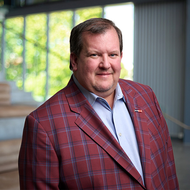 A man with short brown hair and a trimmed beard wears a red plaid blazer over a light blue shirt, standing indoors with large windows and blurred greenery in the background.