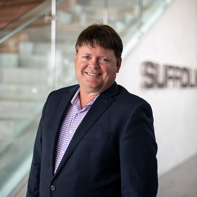A man in a navy blazer and checked shirt, reflecting executive leadership, smiles while standing indoors near a glass staircase, with the word Suffolk visible on a wall in the background.