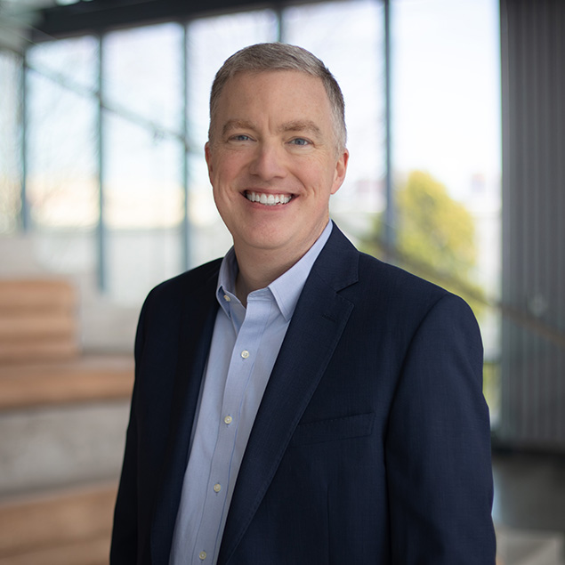A smiling man with short gray hair, exemplifying executive leadership, wears a navy blue suit jacket and light blue shirt as he stands indoors in a modern, well-lit building with large windows.