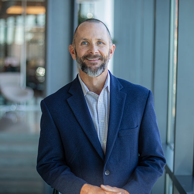 A middle-aged man with a beard and short hair, wearing a blue blazer and a checked shirt, stands indoors by large windows, smiling at the camera. The background is softly blurred office space.