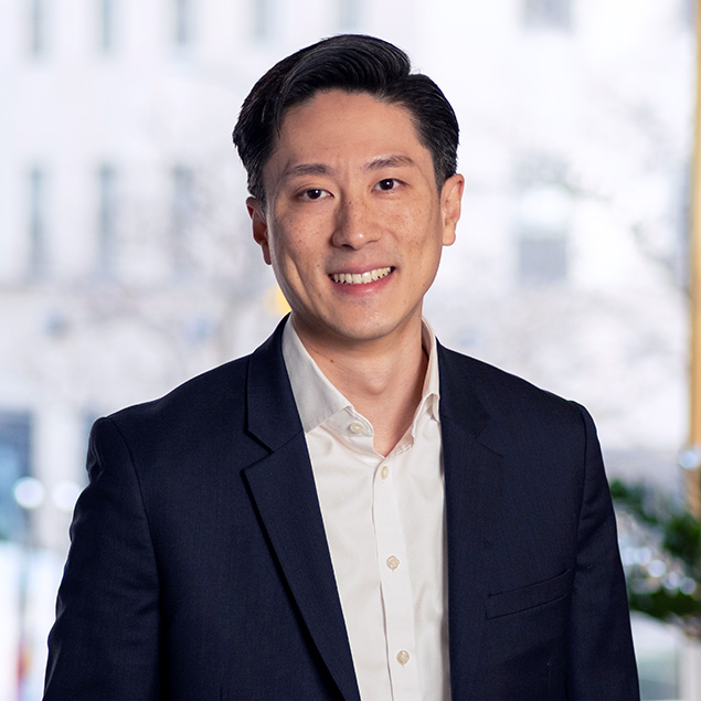 A man with short dark hair, wearing a dark suit jacket and a white dress shirt, stands indoors and smiles at the camera. The background is softly blurred, showing bright windows and out-of-focus elements.