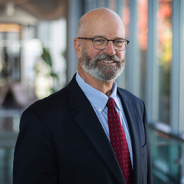 A smiling older man with glasses, a bald head, and a gray beard wears a dark suit, blue striped shirt, and red tie—exuding executive leadership as he stands in a modern hallway filled with natural light.